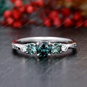 Può includere: Un anello d'argento con tre gemme color teal e pietre bianche più piccole su entrambi i lati. L'anello ha un design a fascia intrecciata. Lo sfondo è sfocato, con sfumature di rosso e giallo.