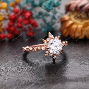 Puede incluir: Un anillo de compromiso de oro rosa con una gran piedra central redonda rodeada de piedras m&aacute;s peque&ntilde;as. El anillo presenta un dise&ntilde;o de estrella con rayos puntiagudos.