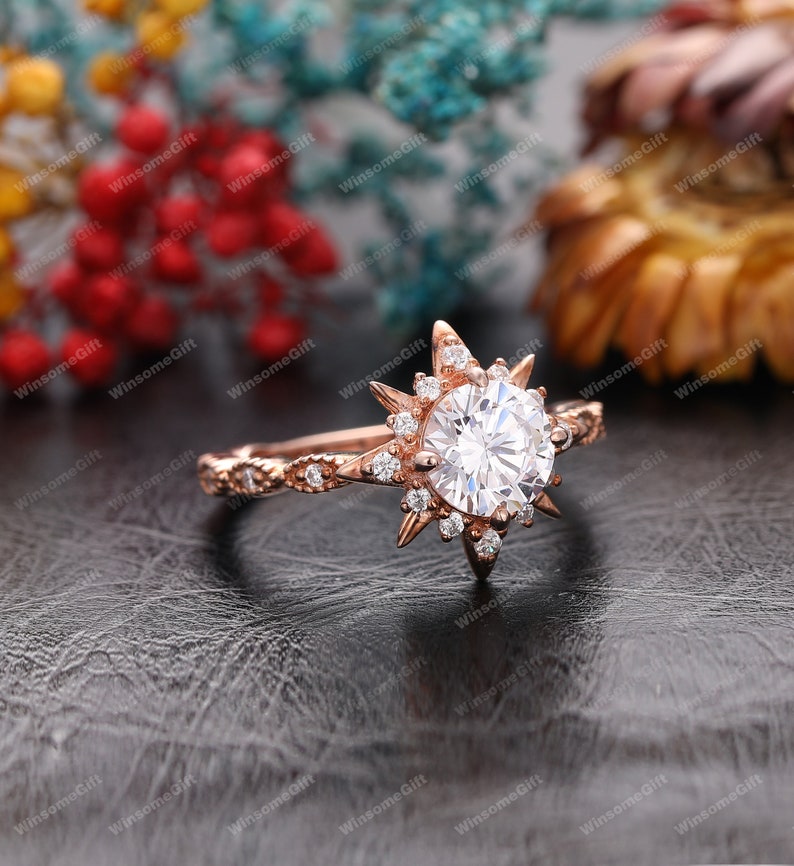 Puede incluir: Un anillo de compromiso de oro rosa con una gran piedra central redonda rodeada de piedras m&aacute;s peque&ntilde;as. El anillo presenta un dise&ntilde;o de estrella con rayos puntiagudos.