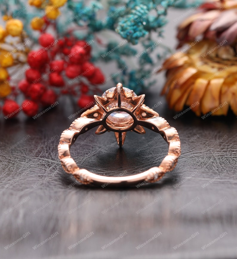 Puede incluir: Un anillo de compromiso de oro rosa con una piedra central grande, redonda y transparente rodeada de piedras m&aacute;s peque&ntilde;as. El anillo tiene un dise&ntilde;o floral delicado con un patr&oacute;n de estrella.