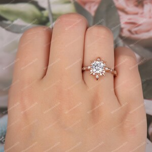 Puede incluir: Un anillo de compromiso de oro rosa con una piedra central grande, redonda y clara rodeada de piedras m&aacute;s peque&ntilde;as en un dise&ntilde;o de estallido de estrellas.