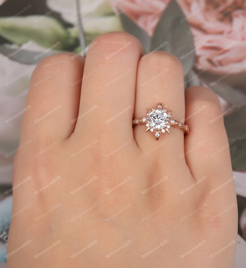 Puede incluir: Un anillo de compromiso de oro rosa con una piedra central grande, redonda y clara rodeada de piedras m&aacute;s peque&ntilde;as en un dise&ntilde;o de estallido de estrellas.