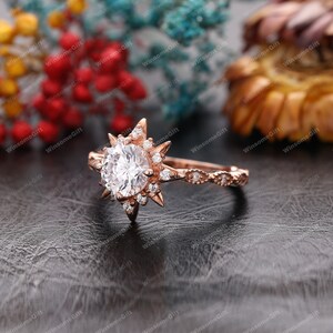 Puede incluir: Anillo de compromiso de oro rosa con una gran piedra central redonda rodeada de piedras m&aacute;s peque&ntilde;as. El anillo presenta un dise&ntilde;o de estrella con rayos puntiagudos.