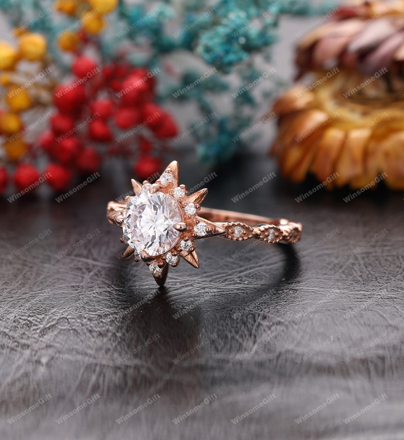 Puede incluir: Anillo de compromiso de oro rosa con una gran piedra central redonda rodeada de piedras m&aacute;s peque&ntilde;as. El anillo presenta un dise&ntilde;o de estrella con rayos puntiagudos.