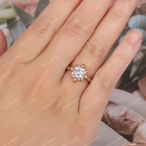 Puede incluir: Un anillo de compromiso de oro rosa con una gran piedra central redonda rodeada de piedras m&aacute;s peque&ntilde;as en un dise&ntilde;o de copo de nieve.