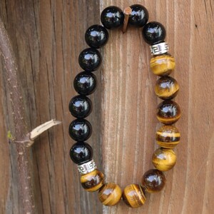 Könnte beinhalten: Ein schwarzes und goldenes Perlenarmband mit silbernen Akzenten. Das Armband besteht aus schwarzen Onyx- und Tigerauge-Perlen.