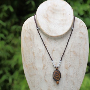 Op de afbeelding: Een ketting van bruin lederen koord met witte kralen en een bruin en oranje hanger. De hanger heeft een geometrisch ontwerp.