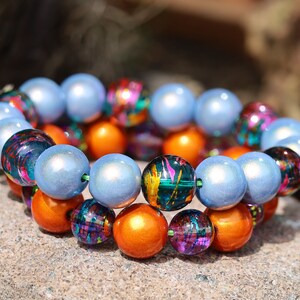 Op de afbeelding: Twee armbanden gemaakt van gekleurde glazen kralen. De ene armband is gemaakt van blauwe kralen en de andere van oranje, paarse en groene kralen.