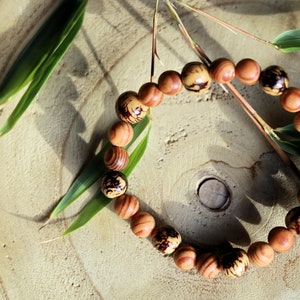 Op de afbeelding: Een houten kralenarmband met bruine en zwarte markeringen op elke kraal. De armband ligt op een lichtbruin houten oppervlak met groene bladeren.