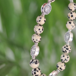 Op de afbeelding: Een armband gemaakt met witte en zwarte gevlekte kralen en zilveren tussenstukken. De tussenstukken hebben een cirkelvormig ontwerp.