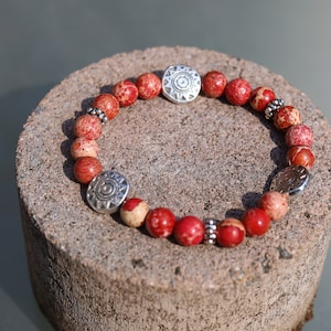 Könnte beinhalten: Armband aus rotem und weißem Jaspis mit silbernen Akzenten. Das Armband besteht aus runden Perlen und zwei größeren silbernen Anhängern mit einem Sonnenstrahl-Design.