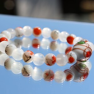 Op de afbeelding: Witte en rode kralen armband met een witte en gouden kraal met rode en groene accenten. De armband ligt op een blauw reflecterend oppervlak.