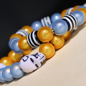 Op de afbeelding: Een armband gemaakt van blauwe, gouden en zwarte kralen. De armband heeft een schedelbedel.