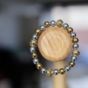 Könnte beinhalten: Ein silbernes und goldenes Perlenarmband auf einem Holzständer. Die Perlen sind rund und glatt, und das Armband ist aus Metall mit einem schlichten Design.