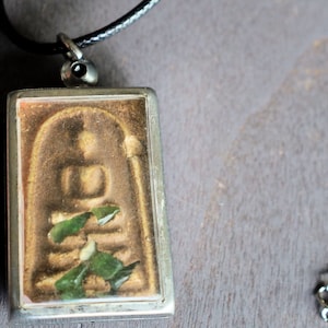 Op de afbeelding: Een rechthoekige hanger met een zilveren frame en een zwart koord. De hanger heeft een goudkleurige afbeelding van een zittende figuur met groene accenten. De achtergrond is een gedempte bruine kleur.