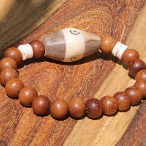 Könnte beinhalten: Ein Holzperlenarmband mit einem weißen Knochenanhänger mit einem kreisförmigen Design. Das Armband ist braun und hat eine natürliche Holzoptik.