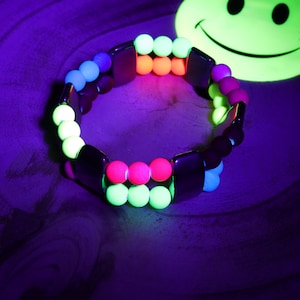 Op de afbeelding: Een veelkleurige glow-in-the-dark kralen armband met zwarte kralen. De armband ligt op een houten oppervlak.