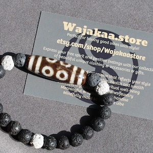 May include: Black lava rock and white bead bracelet with a brown and white Dzi bead charm. The bracelet is on a gray surface with a business card that says "Wajakaa.store" and "etsy.com/shop/Wajakaastore".