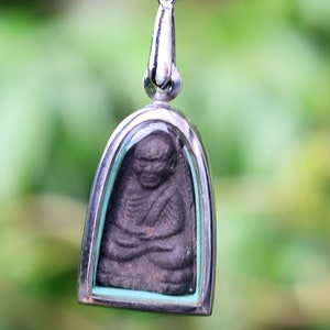 Può includere: Un ciondolo d'argento con una piccola statua di Buddha grigio scuro all'interno. Il ciondolo è su una catena d'argento e ha un bordo blu chiaro attorno alla statua di Buddha.