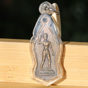 Può includere: Un ciondolo d'argento con una custodia in plastica trasparente, che presenta un'immagine in bronzo di una persona che tiene un'arma. Il ciondolo ha la parola thailandese "พระพิฆเนศ" incisa sul fondo.