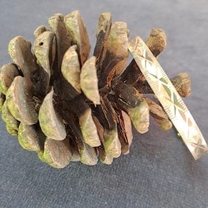May include: A silver-colored bracelet with a geometric star pattern rests next to a brown pine cone. The bracelet is a thin band with a series of etched star and cross designs. The pine cone is brown with green and gray accents.