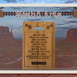 May include: A rectangular bamboo cutting board with a handle, featuring the text "MAY THE SUN...APACHE BLESSING" in black. The board is decorated with floral and bird designs. The background shows a desert landscape with mesas and a blue sky.