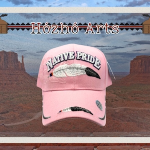 May include: Pink baseball cap with the text "NATIVE PRIDE" and a feather design. The cap also features the text "Hózhó Arts". The background shows a desert landscape with rock formations.