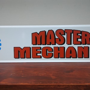 May include: White rectangular sign with a blue and white ASE Certified logo on the left and the text "MASTER MECHANIC" in red letters on the right.