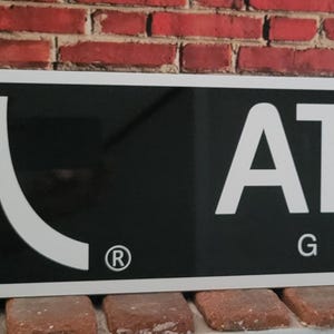 May include: A rectangular black sign with the Atari logo and the word "ATARI GAMES" in white. The sign has a white border and is mounted on a brick wall. The Atari logo is a stylized mountain range.