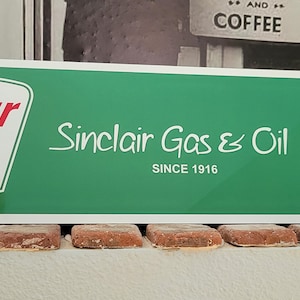 May include: A green and white rectangular sign with the Sinclair logo, a green dinosaur, and the text "Sinclair Gas & Oil Since 1916". The sign also features a white Route 66 shield with the number 66 in black.