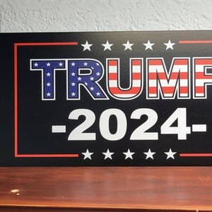 May include: Black and white sign with the text "TRUMP -2024- " in white letters with red and blue stars. The sign also features a sheriff's badge, an American flag, and a snake with the text "Don't Tread On Me". The text "never surrender" is at the bottom of the sign.