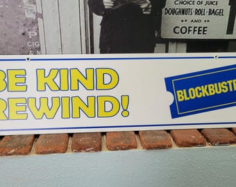 May include: A rectangular white sign with a blue border and the words "BE KIND REWIND!" in yellow. The Blockbuster logo is on the right. The sign is on a brick ledge.