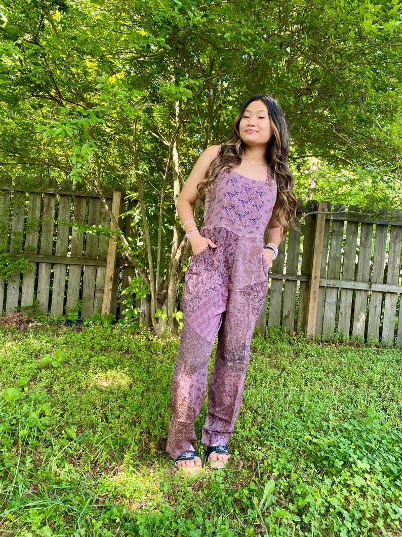 May include: A lavender jumpsuit with a square neckline and pockets. The top has a patterned design, and the trousers feature a patchwork of patterns. The person is wearing sandals and standing in front of a wooden fence and green foliage.