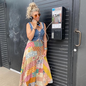 Puede incluir: Un chaleco vaquero con cordones se combina con una falda larga patchwork en varios colores y estampados. La persona está usando una cabina telefónica vintage. La falda presenta una mezcla de diseños florales y geométricos. La persona lleva gafas de sol.