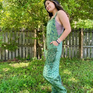 May include: A green floral patterned jumpsuit with wide legs and pockets. The straps are visible over a pink top. The person is wearing sandals with black straps and wooden soles. The background includes a wooden fence and green foliage.