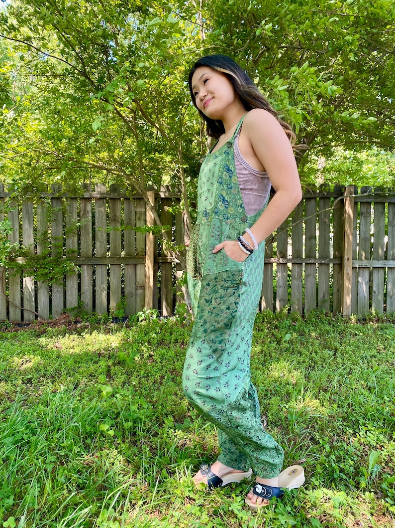May include: A green floral patterned jumpsuit with wide legs and pockets. The straps are visible over a pink top. The person is wearing sandals with black straps and wooden soles. The background includes a wooden fence and green foliage.