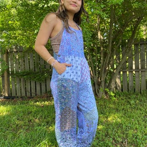 May include: A light blue patchwork jumpsuit with floral and geometric patterns. The jumpsuit features adjustable straps, pockets, and a relaxed fit. The model is wearing a brown vest top underneath. The jumpsuit is made of a lightweight fabric.