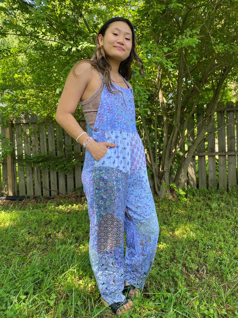 May include: A light blue patchwork jumpsuit with floral and geometric patterns. The jumpsuit features adjustable straps, pockets, and a relaxed fit. The model is wearing a brown vest top underneath. The jumpsuit is made of a lightweight fabric.