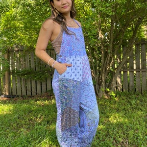 May include: A light blue jumpsuit with a patchwork design featuring floral and geometric patterns. The jumpsuit has a bib-style top with adjustable straps and pockets. The model is wearing a brown top underneath. The background includes green grass and trees.