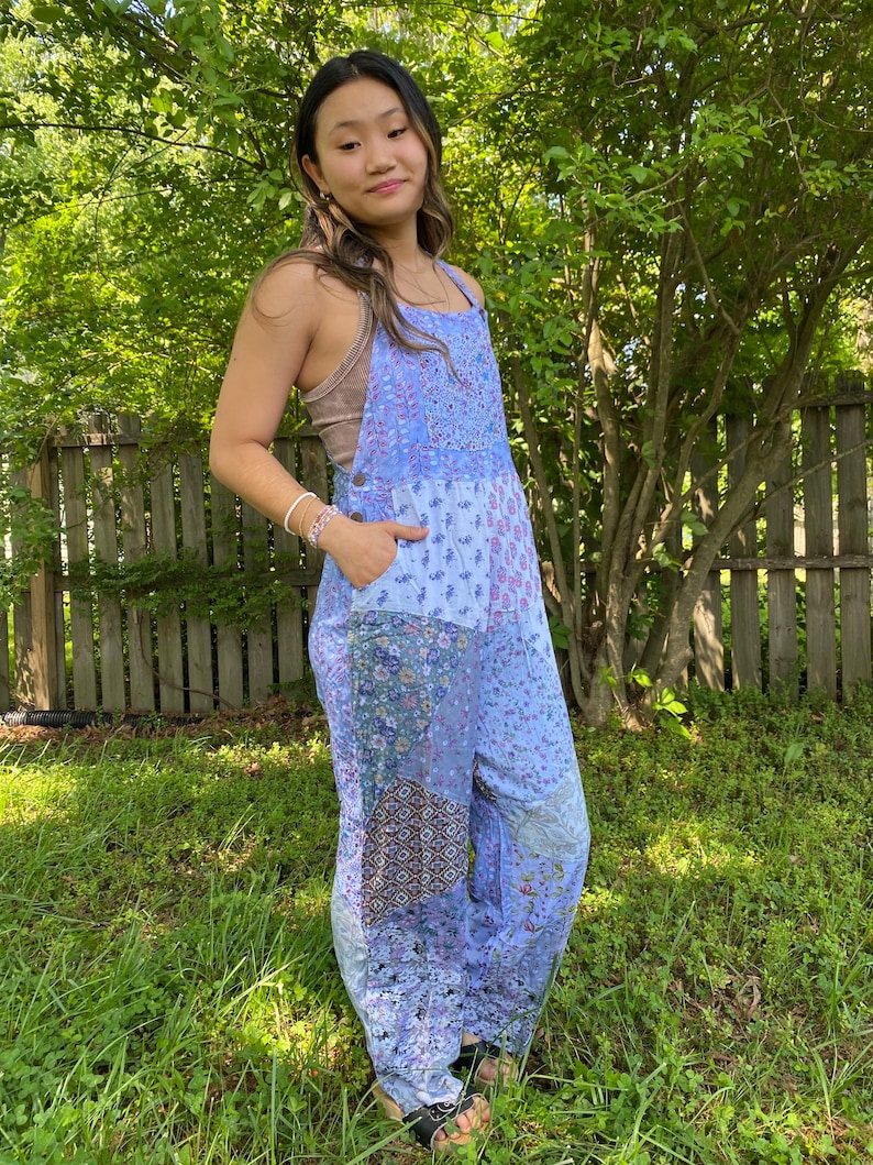 May include: A light blue jumpsuit with a patchwork design featuring floral and geometric patterns. The jumpsuit has a bib-style top with adjustable straps and pockets. The model is wearing a brown top underneath. The background includes green grass and trees.