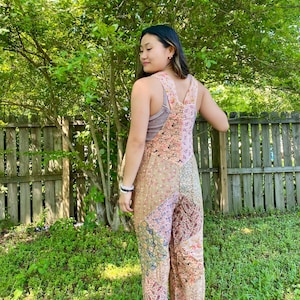 May include: A patchwork jumpsuit in shades of pink, beige, and green. The jumpsuit features a floral and paisley pattern, with wide legs and adjustable straps. The model is standing outside in front of a wooden fence and green foliage.