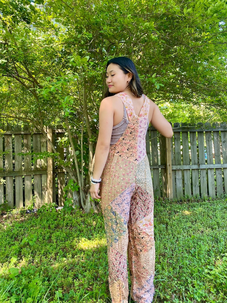 May include: A patchwork jumpsuit in shades of pink, beige, and green. The jumpsuit features a floral and paisley pattern, with wide legs and adjustable straps. The model is standing outside in front of a wooden fence and green foliage.