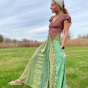 May include: A woman wearing a brown top and a long, flowing skirt with a patchwork design of green and floral patterns. The skirt has a high waist and a slit. The woman is wearing sandals and a headscarf, standing in a grassy field.
