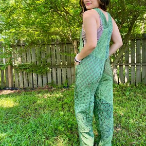May include: Green patchwork jumpsuit featuring floral and paisley patterns. The jumpsuit has a bib-style top with adjustable straps and wide legs. The model is wearing sandals. The background includes a wooden fence and green foliage.