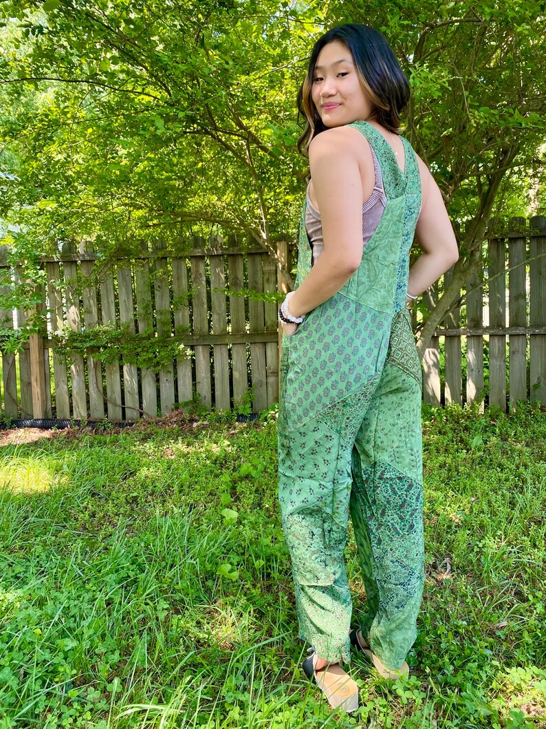 May include: Green patchwork jumpsuit featuring floral and paisley patterns. The jumpsuit has a bib-style top with adjustable straps and wide legs. The model is wearing sandals. The background includes a wooden fence and green foliage.