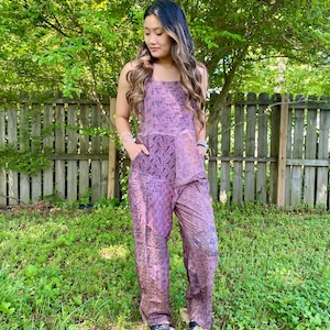 May include: A lavender jumpsuit with a floral pattern. The jumpsuit has a square neckline, wide legs, and pockets. The model is wearing sandals. The background includes a wooden fence and green foliage.