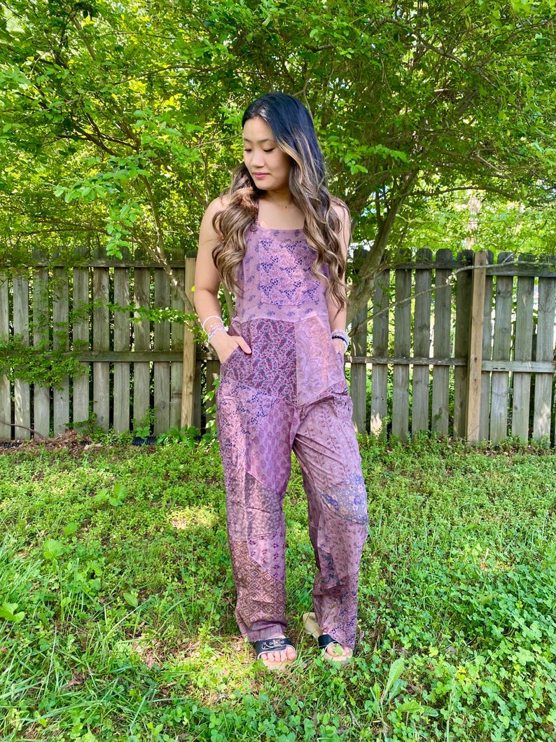 May include: A lavender jumpsuit with a floral pattern. The jumpsuit has a square neckline, wide legs, and pockets. The model is wearing sandals. The background includes a wooden fence and green foliage.