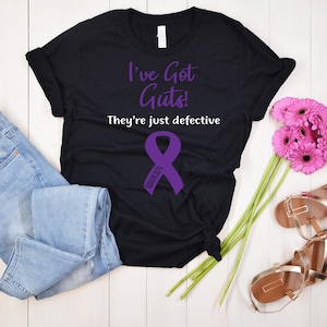 May include: Black t-shirt with the text "I've Got Guts! They're just defective" and a purple ribbon with the word "Defective" on it.