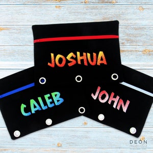 May include: Three black pencil cases with colorful embroidered names: Joshua, Caleb, and John. Each case has a zipper closure and metal grommets.