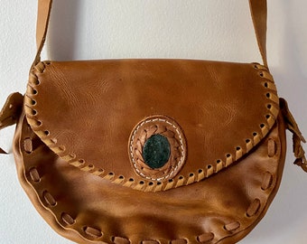May include: A brown leather crossbody bag with a decorative stitched design and a green stone accent.